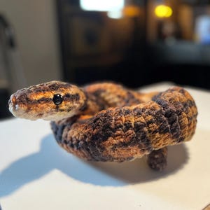 Op de afbeelding: Gehaakte slang in bruine, zwarte en oranje tinten. Het handgemaakte reptiel is opgerold, met een gedetailleerde kop en zwarte ogen. Dit zachte beeldhouwwerk is een uniek decoratie-artikel.