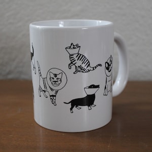 May include: White ceramic mug with black illustrations of cats and dogs wearing cone collars. The mug has a handle on the right side.