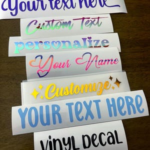 Può includere: Una collezione di decalcomanie in vinile con varie opzioni di testo in diversi colori e caratteri. Le decalcomanie includono frasi come "Your text here", "Custom Text" e "Personalize". La decalcomania in basso recita "Vinyl Decal".