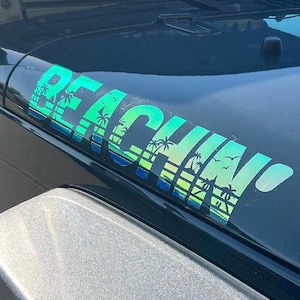 Puede incluir: Primer plano del capó negro de un vehículo con una calcomanía "BEACHIN". La calcomanía presenta un degradado de azul y verde con siluetas de palmeras y pájaros. El capó es ligeramente reflectante.