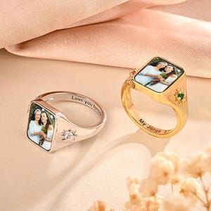 Op de afbeelding: Twee gepersonaliseerde zegelringen, één zilverkleurig en één goudkleurig, elk met een foto in een vierkante setting. De zilveren ring heeft de inscriptie "Love you forever". De gouden ring heeft de inscriptie "My Jorge". Beide ringen hebben decoratieve accenten.