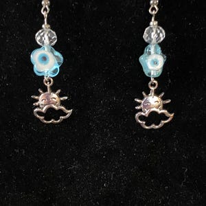 May include: A pair of dangle earrings with silver-tone hooks. Each earring features clear beads, a light blue flower-shaped bead with a white and blue eye design, and a silver-tone sun and cloud charm. The earrings are set against a black background.
