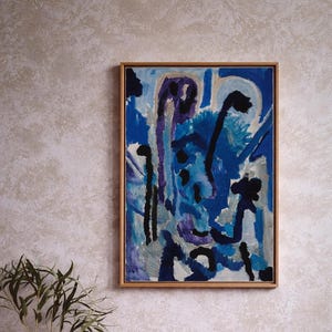 Può includere: Dipinto astratto in una cornice di legno, con sfumature di blu, viola e nero. L'opera d'arte ha uno stile moderno ed espressivo. Un vaso bianco dal design unico e una piccola tazza beige sono in primo piano.