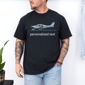 Op de afbeelding: Een zwart T-shirt met korte mouwen voorzien van een afbeelding van een lichtblauw en wit eenmotorig vliegtuig. Onder de illustratie van het vliegtuig staan de woorden "personalized text" in witte, schreefloze kleine letters op de borst.