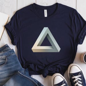 May include: Navy blue t-shirt featuring a 3D impossible triangle design in shades of white and light green. The shirt is paired with blue jeans and navy blue sneakers with white soles. A casual, everyday wear item.