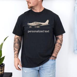 Può includere: Una t-shirt nera a maniche corte con un'illustrazione grafica di un aereo a elica di colore chiaro visto di profilo. Sotto la grafica dell'aereo, le parole "personalized text" sono stampate in un semplice carattere bianco senza grazie.