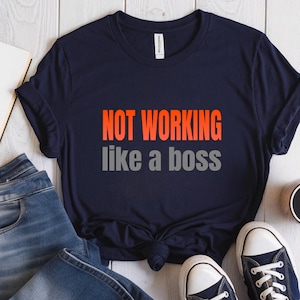 May include: Navy blue t-shirt with the text "NOT WORKING like a boss" in orange and gray. The shirt is styled with blue jeans and white-soled sneakers.