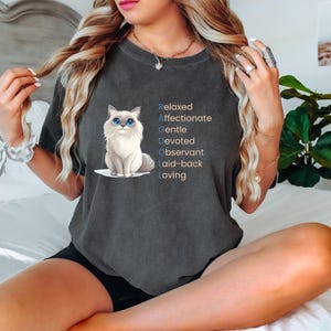 Op de afbeelding: Donkergrijs t-shirt met een Ragdoll kat illustratie met blauwe ogen. Het shirt heeft het woord "RAGDOLL" in blauwe en bruine tekst, met beschrijvende woorden voor elke letter: Relaxed, Affectionate, Gentle, Devoted, Observant, Laid-back en Loving.