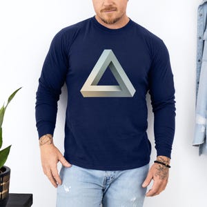 May include: Navy blue long-sleeve shirt featuring a 3D impossible triangle graphic in shades of gray and beige. The shirt is made of a soft material and is suitable for casual wear. The shirt is paired with blue jeans.