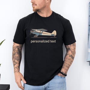 Op de afbeelding: Zwart t-shirt met een gedetailleerde illustratie van een vintage vliegtuig in blauwe, crème- en bruine tinten. De tekst "personalized text" is onder het vliegtuig gedrukt. Het shirt is gemaakt van een zacht materiaal.