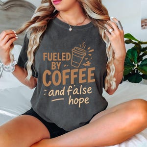May include: Dark gray t-shirt with the text "Fueled by Coffee and false hope" in brown lettering. The design includes a coffee cup graphic. The shirt is a casual wear item.