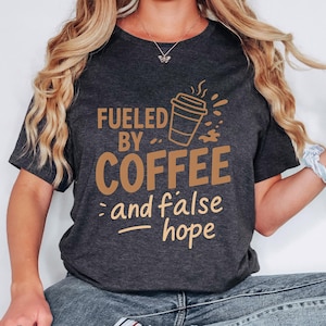 May include: Dark gray t-shirt with the text "Fueled by Coffee and false hope" in brown lettering. A coffee cup graphic is also featured. The shirt is a casual wear item.