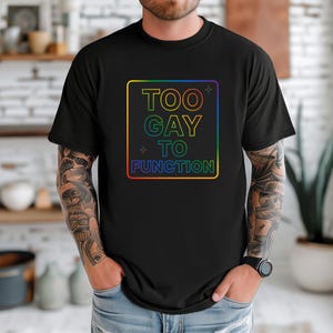 Peut inclure: T-shirt noir avec un motif carré aux couleurs de l'arc-en-ciel. Le motif comporte le texte "TOO GAY TO FUNCTION" dans une police dégradée arc-en-ciel. Le t-shirt est à col rond et à manches courtes.