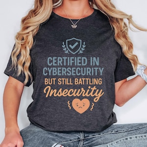 May include: Dark grey t-shirt featuring the text "CERTIFIED IN CYBERSECURITY BUT STILL BATTLING Insecurity" in blue and orange. The design includes a shield with a checkmark and a sad heart graphic. The shirt has a crew neck.