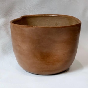 May include: A handmade, brown ceramic bowl with a unique, asymmetrical shape. The bowl has a smooth, unglazed exterior and a slightly lighter interior. The rim features a subtle indentation. This pottery piece is suitable for serving or display.