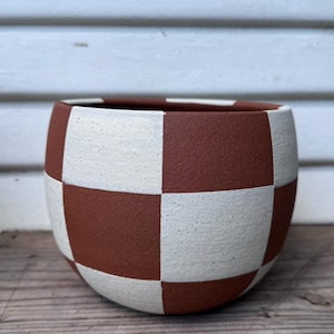 May include: A ceramic planter with a checkerboard pattern in terracotta and white. The round, low-profile pot sits on a wooden surface. The planter is suitable for indoor or outdoor use.