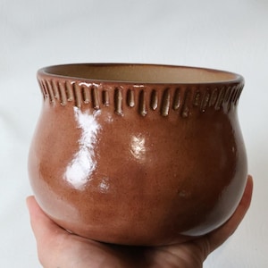 May include: A brown ceramic bowl with a glossy finish. The rim features a decorative, textured pattern. The bowl is held in a hand, showcasing its rounded shape and earthy tones. A simple, yet elegant piece for home decor.