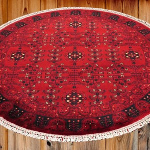 May include: Oval red rug with intricate geometric patterns and a white fringe border. The rug's design features black and gold accents, creating a traditional aesthetic. The rug is placed on a wooden surface.