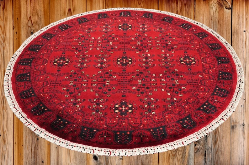 May include: Oval red rug with intricate geometric patterns and a white fringe border. The rug's design features black and gold accents, creating a traditional aesthetic. The rug is placed on a wooden surface.