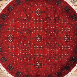 May include: A round, red rug with an intricate geometric pattern. The rug features a central medallion and repeating motifs in black, gold, and navy blue. The rug has a white fringe border and is displayed on a wooden surface.