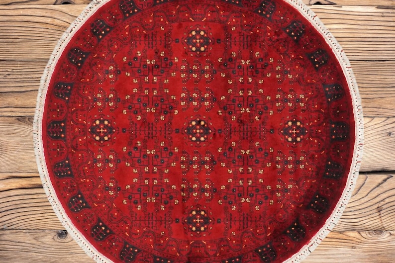 May include: A round, red rug with an intricate geometric pattern. The rug features a central medallion and repeating motifs in black, gold, and navy blue. The rug has a white fringe border and is displayed on a wooden surface.
