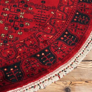 May include: A round, red rug with intricate black and white patterns. The rug features a detailed border and a fringed edge. The design is a traditional style, with a focus on geometric shapes and floral motifs. The rug is placed on a wooden surface.