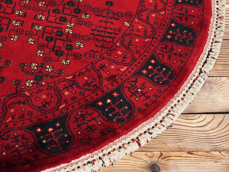 May include: A round, red rug with intricate black and white patterns. The rug features a detailed border and a fringed edge. The design is a traditional style, with a focus on geometric shapes and floral motifs. The rug is placed on a wooden surface.