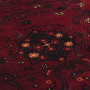 May include: Close-up of a red rug with a detailed pattern. The rug features a central black design with orange and yellow accents, surrounded by smaller floral motifs. The texture appears soft and plush.