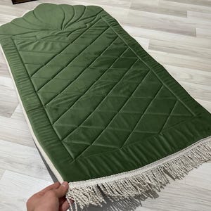 May include: Green prayer rug with a quilted diamond pattern and a decorative scalloped top. The rug has a cream-colored border and fringe. The rug is made of a soft, velvety material.