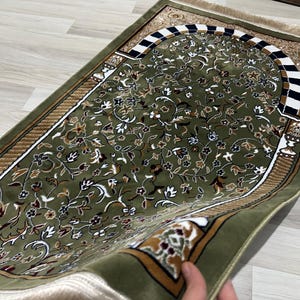May include: Green prayer mat with a floral pattern and an arched border. The mat features a detailed design with white, brown, and gold accents. The rectangular mat has a fringed edge.
