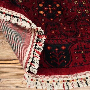 May include: A rolled-up, red oriental rug with a black border and intricate floral patterns. The rug has a cream-colored fringe with black and red accents. The rug is displayed on a wooden surface.