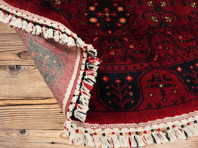 May include: A rolled-up, red oriental rug with a black border and intricate floral patterns. The rug has a cream-colored fringe with black and red accents. The rug is displayed on a wooden surface.
