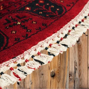 May include: Close-up of a red and black patterned rug with a cream-colored fringe. The rug features a detailed geometric design with a border of cream-colored tassels. The rug is placed on a wooden surface.