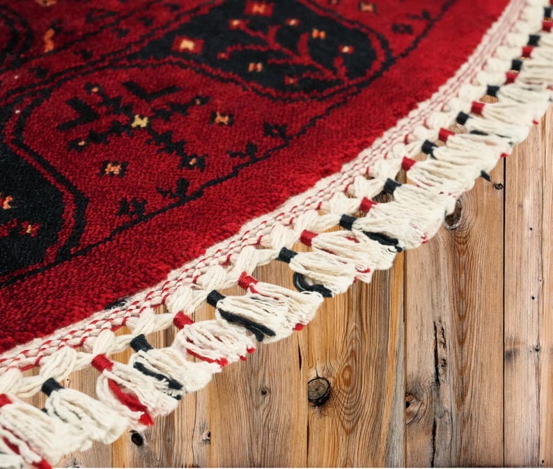 May include: Close-up of a red and black patterned rug with a cream-colored fringe. The rug features a detailed geometric design with a border of cream-colored tassels. The rug is placed on a wooden surface.