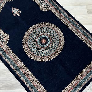 May include: A navy blue prayer rug with an ornate, circular central design in shades of beige, teal, and red. The rug features a decorative border and a small, stylized floral design near the top.