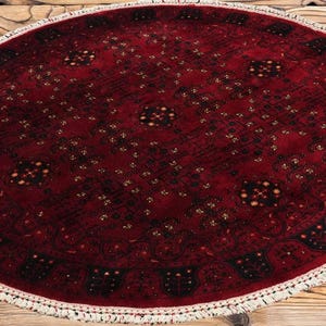 May include: Oval, hand-knotted rug with a deep red field and intricate geometric patterns in black and gold. The rug has a white fringe border. A traditional, decorative floor covering.