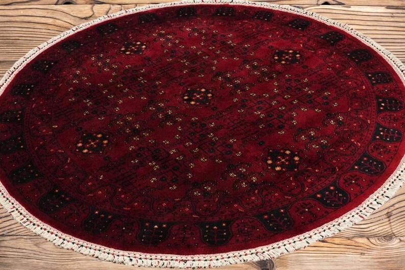 May include: Oval, hand-knotted rug with a deep red field and intricate geometric patterns in black and gold. The rug has a white fringe border. A traditional, decorative floor covering.