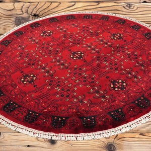 May include: A round, red rug with an intricate geometric pattern and a white fringe. The rug's design features black and cream-colored accents. The rug is placed on a wooden surface.