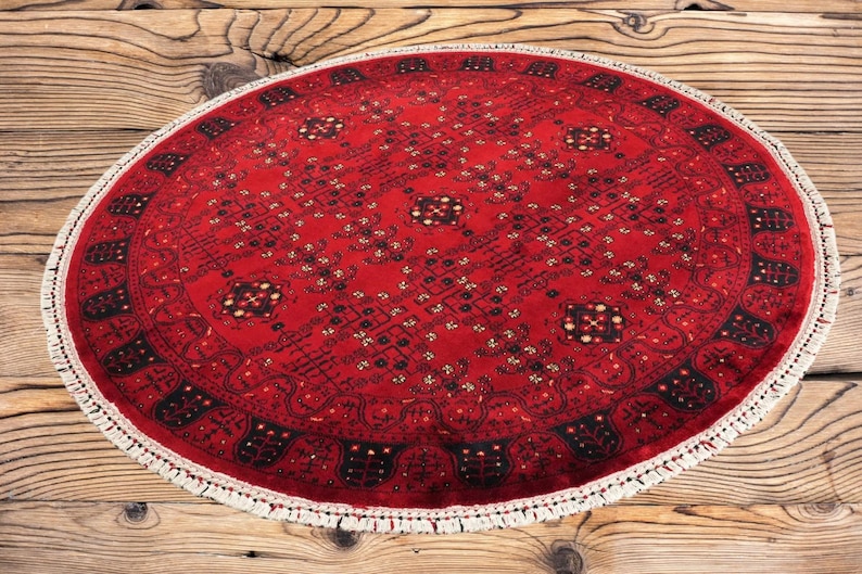 May include: A round, red rug with an intricate geometric pattern and a white fringe. The rug's design features black and cream-colored accents. The rug is placed on a wooden surface.