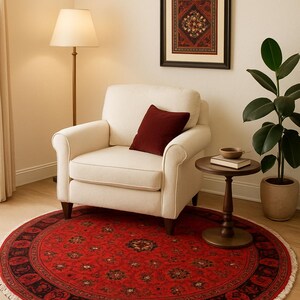May include: A round red rug with an intricate floral pattern and a black border. The rug is placed in a room with a cream-colored armchair, a side table, and a framed artwork. The rug's diameter is approximately 60 inches.
