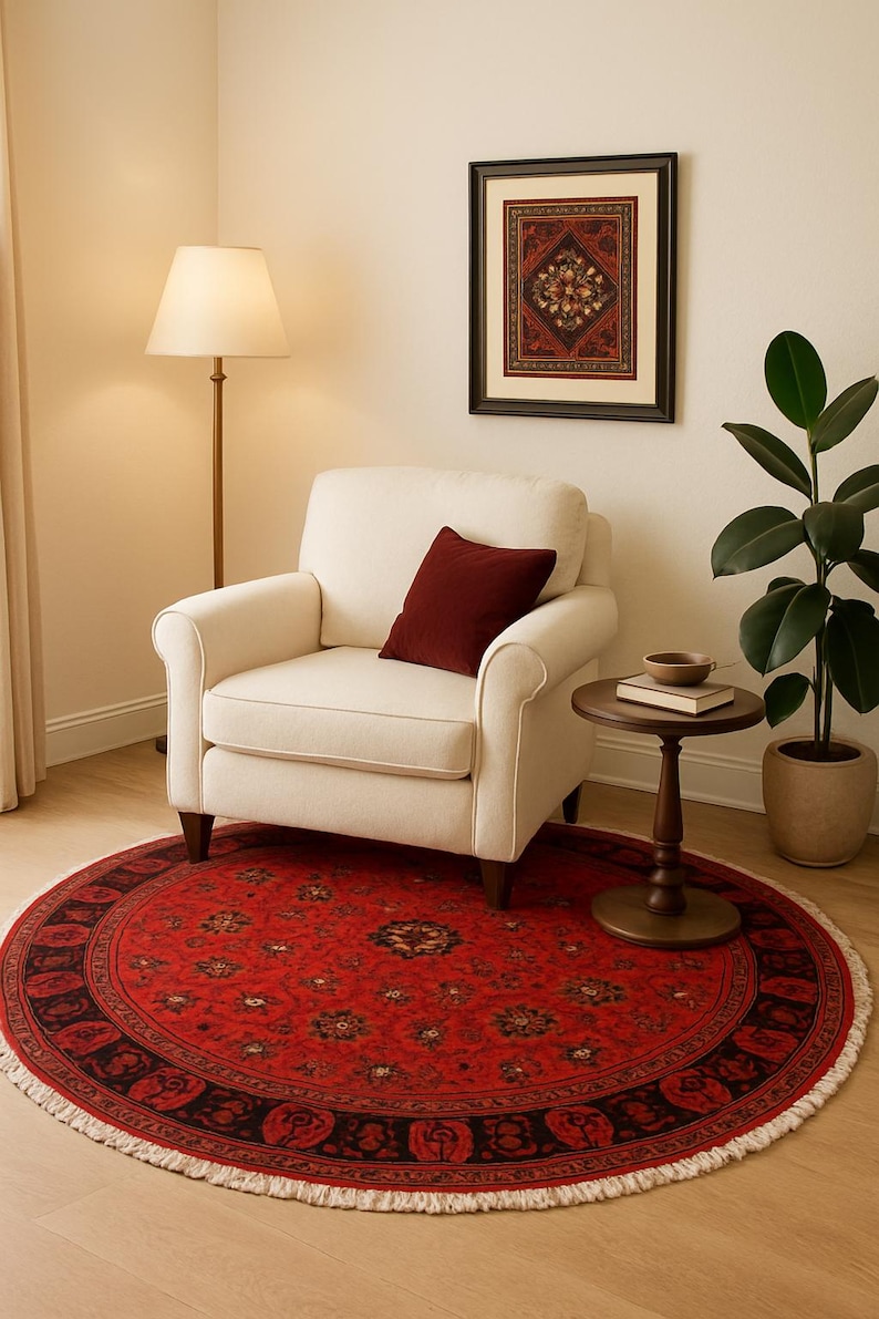 May include: A round red rug with an intricate floral pattern and a black border. The rug is placed in a room with a cream-colored armchair, a side table, and a framed artwork. The rug's diameter is approximately 60 inches.
