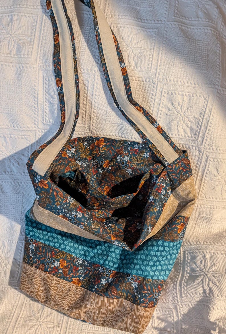May include: A teal blue fabric tote bag with a floral print and brown and beige accents. The bag has a long strap and a patchwork design.