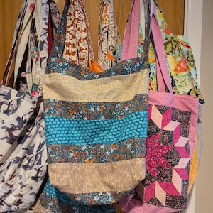 Könnte beinhalten: Eine Sammlung von sieben bunten Tragetaschen, die an Haken hängen. Die Taschen sind aus Stoff mit verschiedenen Mustern, darunter Streifen, Blumenmuster und geometrische Designs.