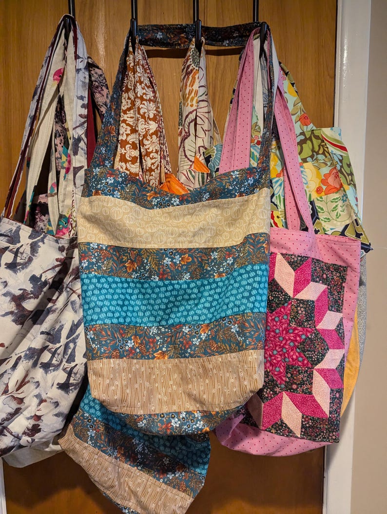 May include: A collection of seven colourful tote bags hanging on hooks. The bags are made of fabric with different patterns, including stripes, floral prints, and geometric designs.