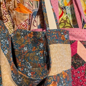 May include: A tote bag made from patchwork fabric with a variety of autumn-themed prints, including pumpkins, leaves, and flowers. The bag has a teal blue base with a tan pocket and a white strap.