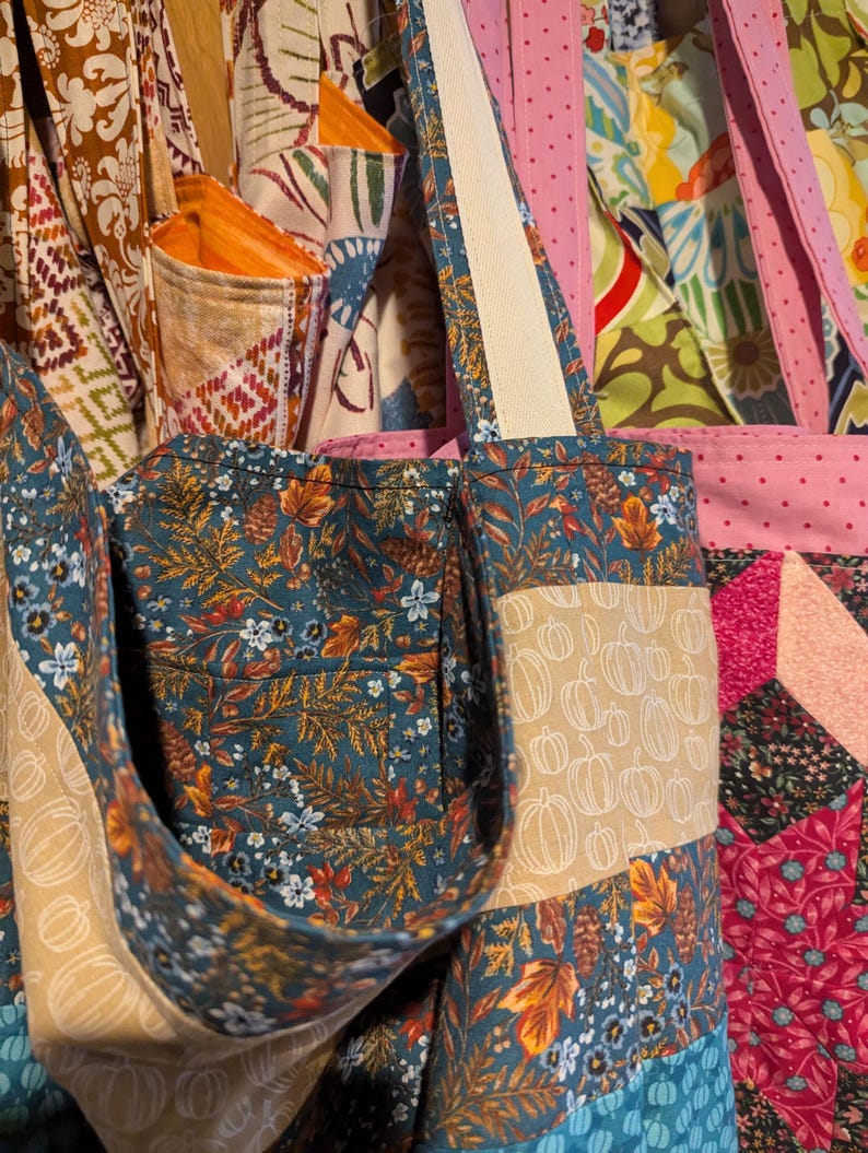May include: A tote bag made from patchwork fabric with a variety of autumn-themed prints, including pumpkins, leaves, and flowers. The bag has a teal blue base with a tan pocket and a white strap.