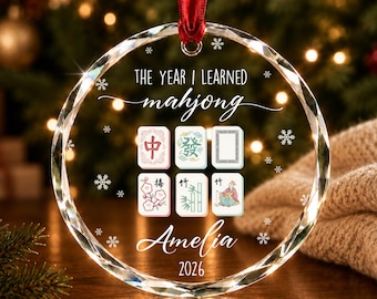 May include: A circular clear glass ornament with faceted edges, hanging from a red ribbon. It features the text "THE YEAR I LEARNED mahjong", the name "Amelia", the year "2026", six mahjong tile illustrations, and small white snowflake accents.