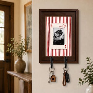 May include: A dark brown wooden wall-mounted key holder featuring a central vertical red and white striped background. An ace of hearts playing card graphic displays a black and white photo of a couple with the text How lucky are we. Two black metal hooks hold two sets of keys.