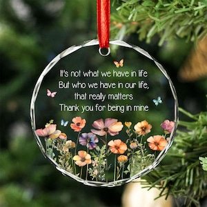 Op de afbeelding: Een rond ornament van helder glas met een gefacetteerde rand, hangend aan een rood lint. Het is voorzien van een bloemmotief met kleurrijke wilde bloemen en vlinders, en de tekst: It's not what we have in life, But who we have in our life, that really matters, Thank you for being in mine.