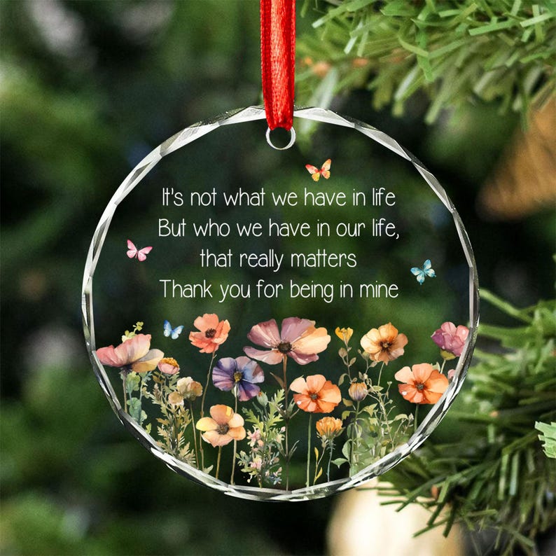 Op de afbeelding: Een rond ornament van helder glas met een gefacetteerde rand, hangend aan een rood lint. Het is voorzien van een bloemmotief met kleurrijke wilde bloemen en vlinders, en de tekst: It's not what we have in life, But who we have in our life, that really matters, Thank you for being in mine.